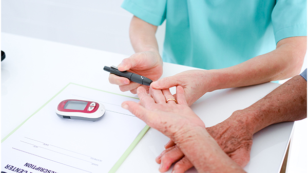 A healthcare professional uses a lancing device on an older person's fingertip near a glucose meter on a white table.